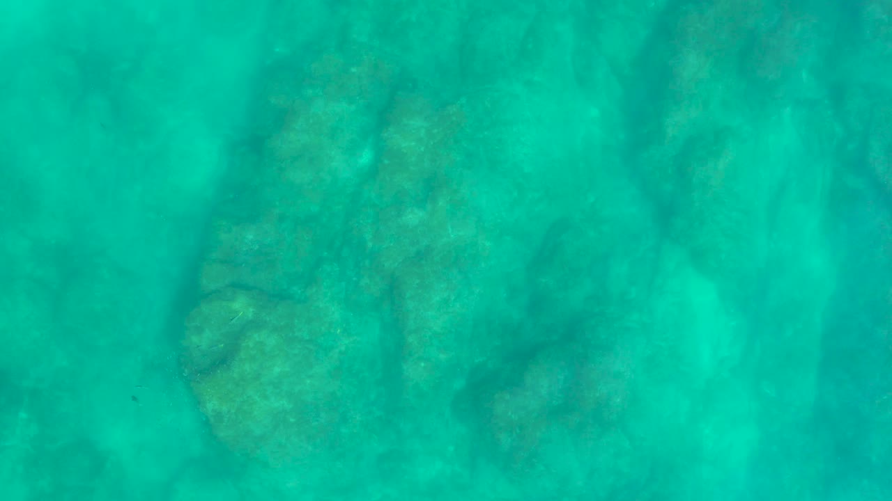 Underwater frame from Baja California Sur showing darker rock forms under clear turquoise water, with no visible people or surface objects.