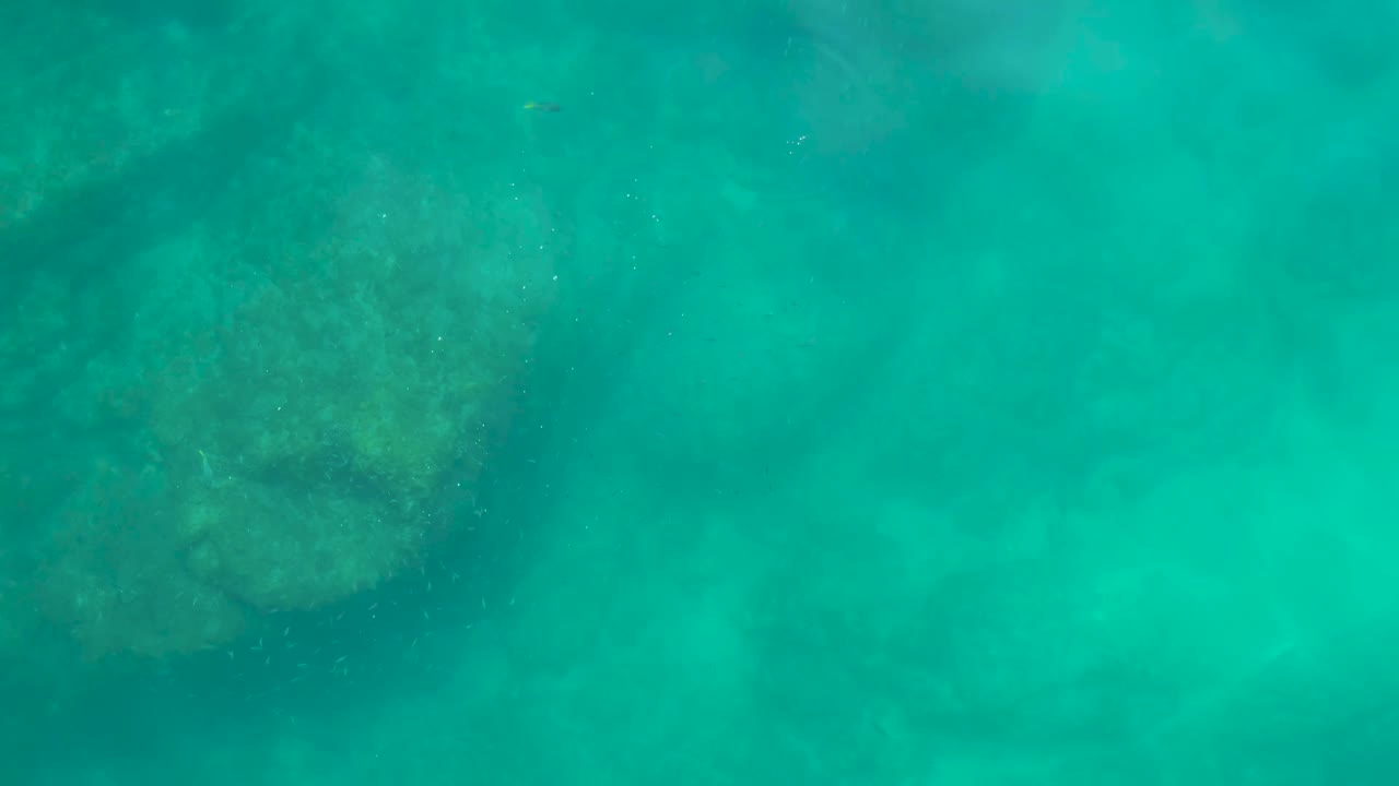 Underwater frame at Shipwrecks Los Cabos with a large muted rock shape left of center beneath turquoise water and fine particles in the water, with no visible people.