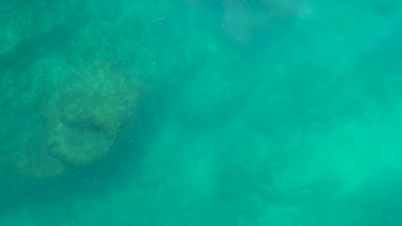 Mid-clip underwater frame at Shipwrecks Los Cabos showing teal water, shadowed rock forms, and a pale sandy patch in the lower frame, with no visible people.