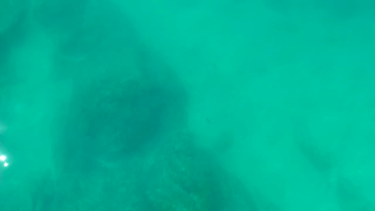 Turquoise underwater view at Shipwrecks Los Cabos with mottled rocky seabed texture spreading across the lower portion and no visible people.