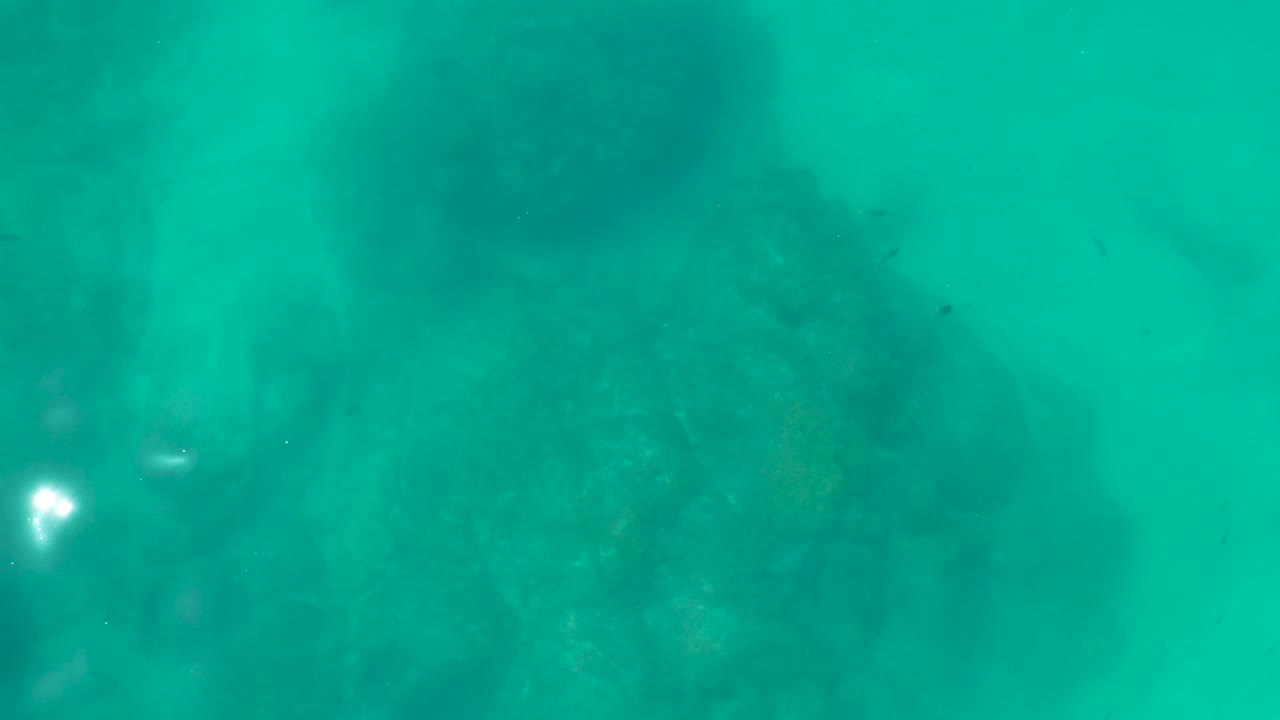 A darker patch of rocky seabed sits beneath calm turquoise water at Shipwrecks Los Cabos, with tiny fish shapes and no visible people.