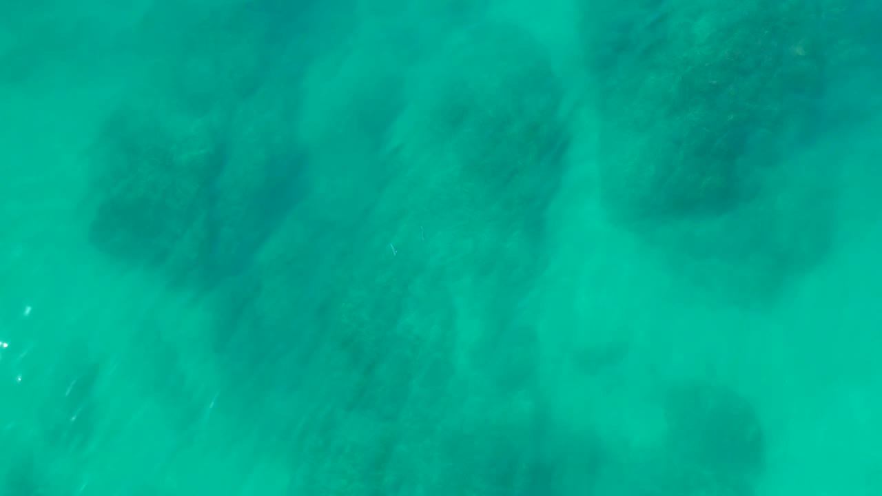 Overhead view of turquoise water at Shipwrecks Los Cabos with dark underwater rock formations, pale sandy patches, soft surface texture, and no visible people.