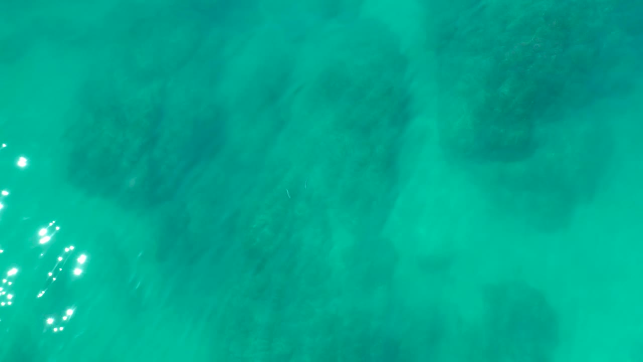 Bright surface glints appear on turquoise water at Shipwrecks Los Cabos while submerged rock formations remain visible below, with no visible people.