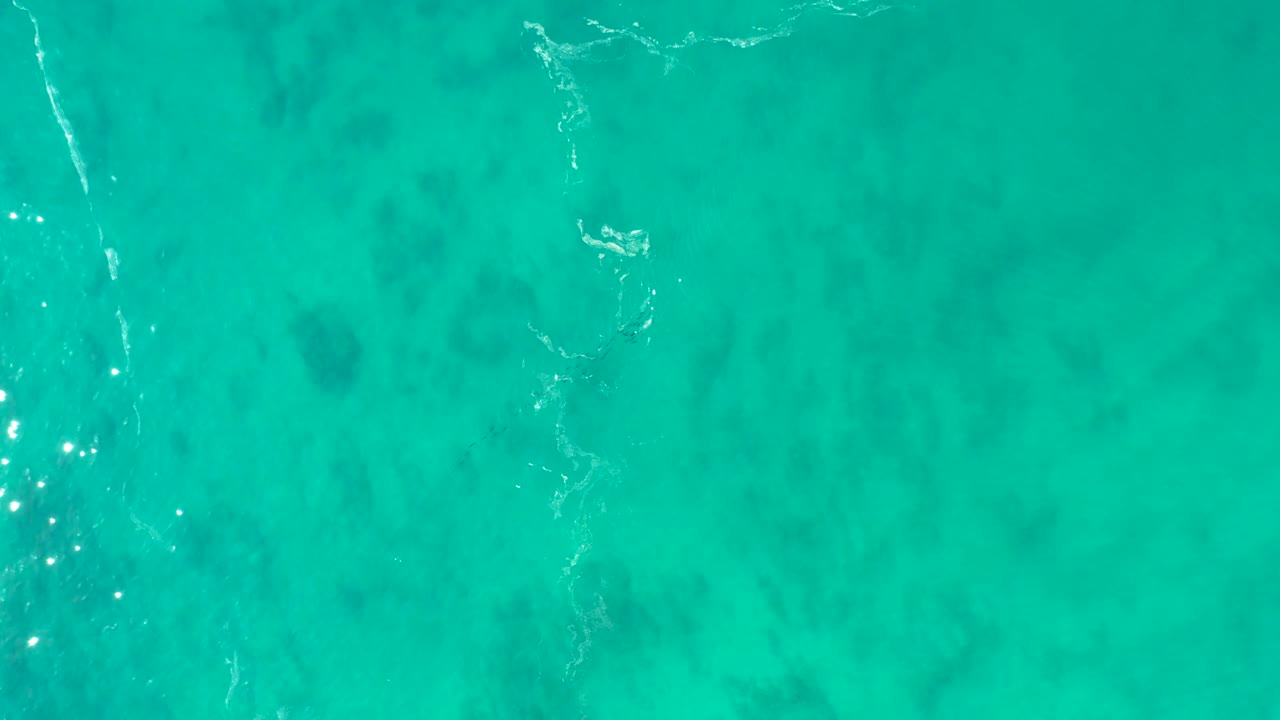 Top-down view of turquoise ocean at Shipwrecks Los Cabos with pale foam streaks near the upper left, darker seabed patches below the surface, and no visible people.