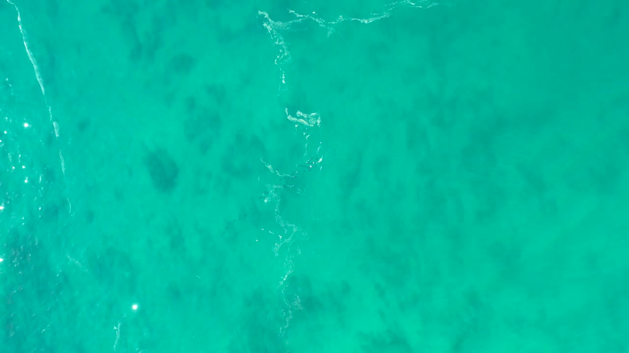 Turquoise shallow ocean at Shipwrecks Los Cabos shows soft darker seabed shapes, a broken white foam mark near the center left, and no visible people.