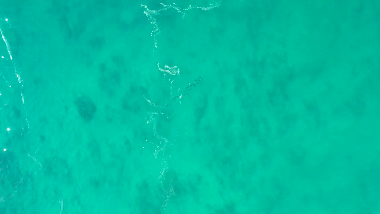 Clear turquoise water fills the frame at Shipwrecks Los Cabos, with scattered dark seabed marks and a narrow foam ripple crossing near the upper left, no visible people.