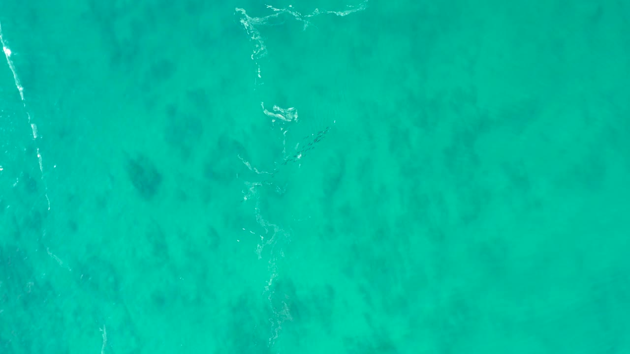 Aerial view over Shipwrecks Los Cabos turquoise sea, showing a pale foam line angled through the frame and soft seabed shadows below, no visible people.