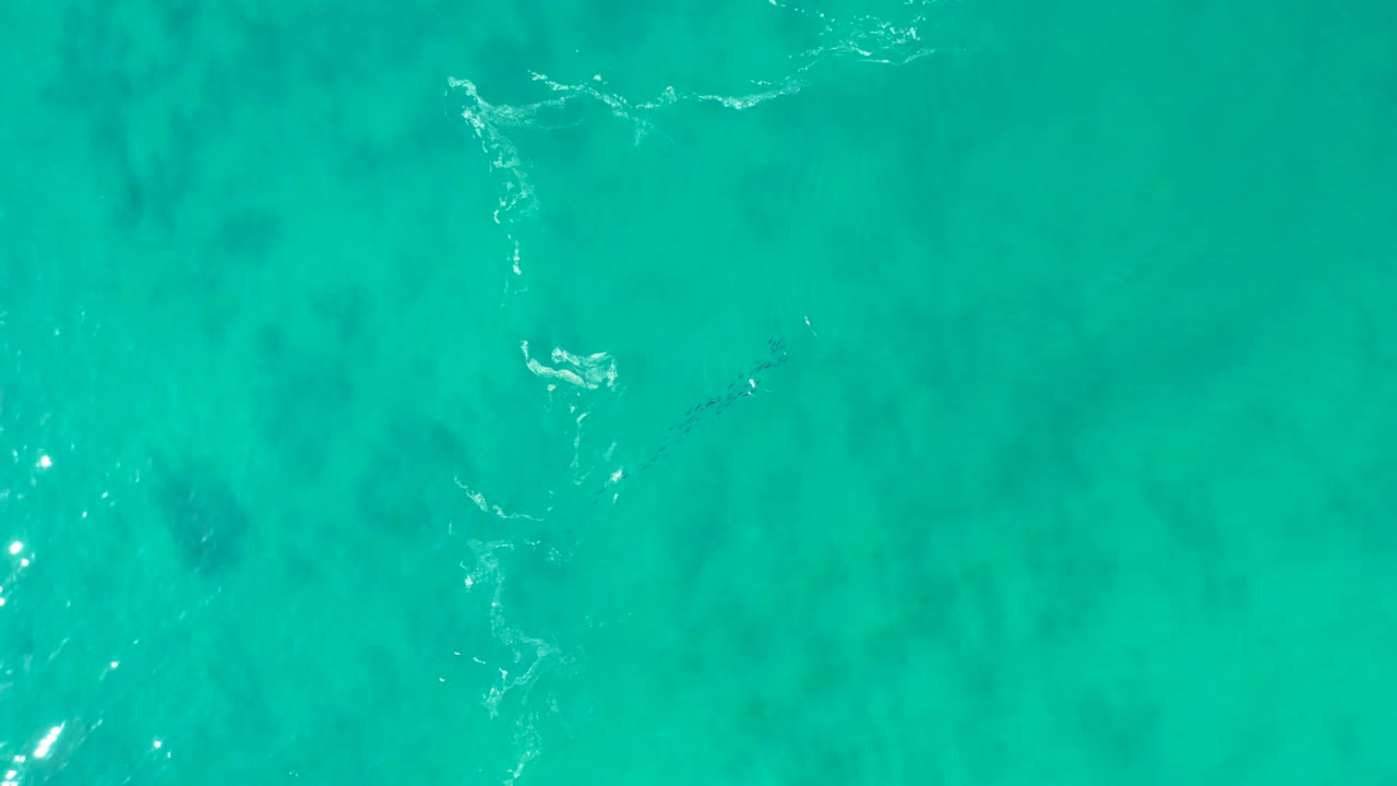 Aerial-looking frame of shallow turquoise water at Shipwrecks Los Cabos with bright sun glints, subtle foam, and darker underwater patches, no visible people.