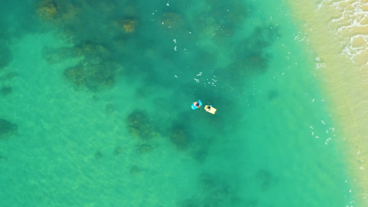 Overhead frame at Shipwrecks Los Cabos shows two people on small bright inflatables in clear turquoise water, with pale sand and thin wave foam along the right edge.