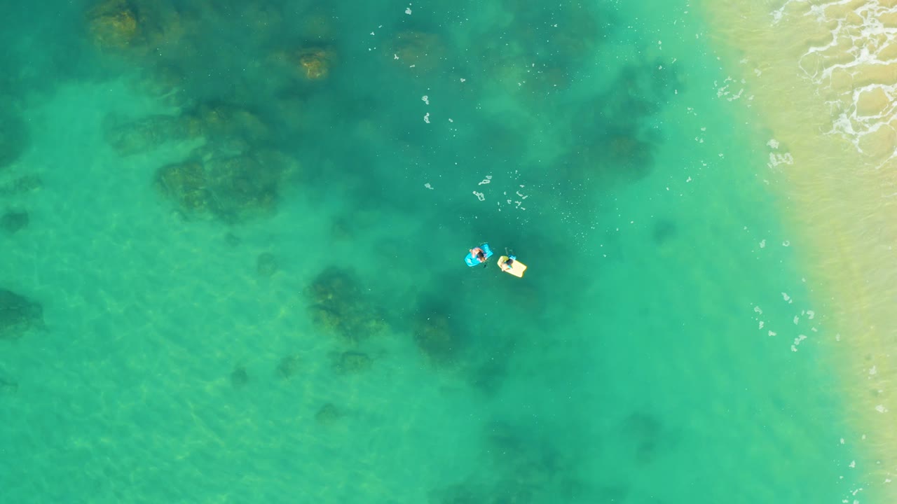 Two small floaters in shallow turquoise ocean beside pale sand.