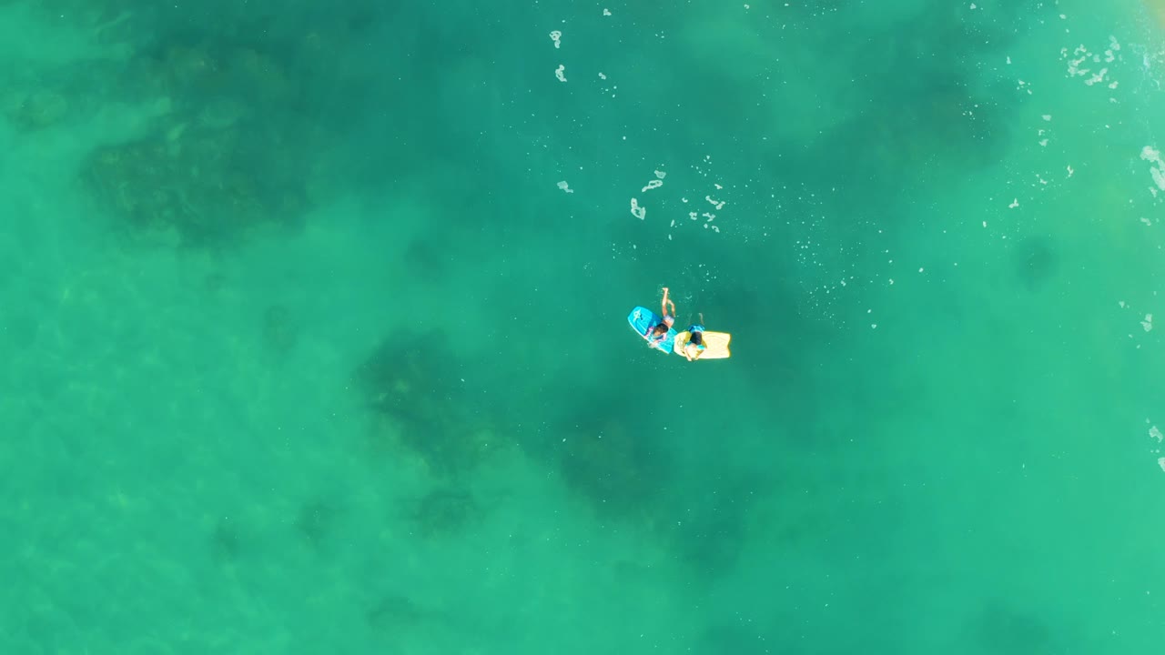 Overhead frame from Shipwrecks Los Cabos showing a person on a small float near the upper-left area of clear turquoise water with dark underwater patches below.