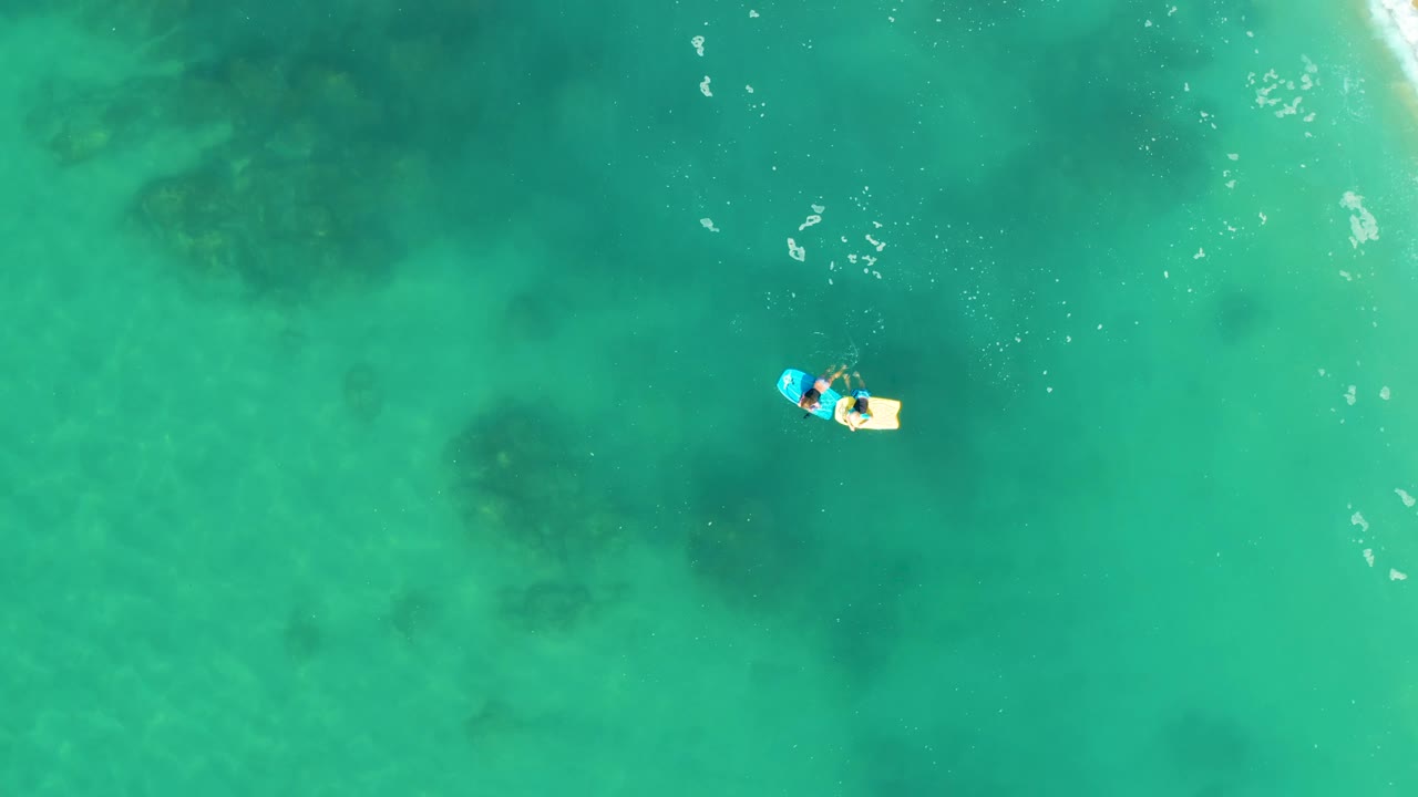 A high overhead frame shows the person on the float near the upper-right side of the water field, with dark submerged patches visible through the turquoise ocean.