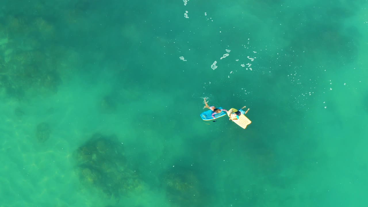 At 0:02, the blue and yellow boards sit toward the upper right of the overhead frame, with the people visible against clear green-blue water at Shipwrecks Los Cabos.