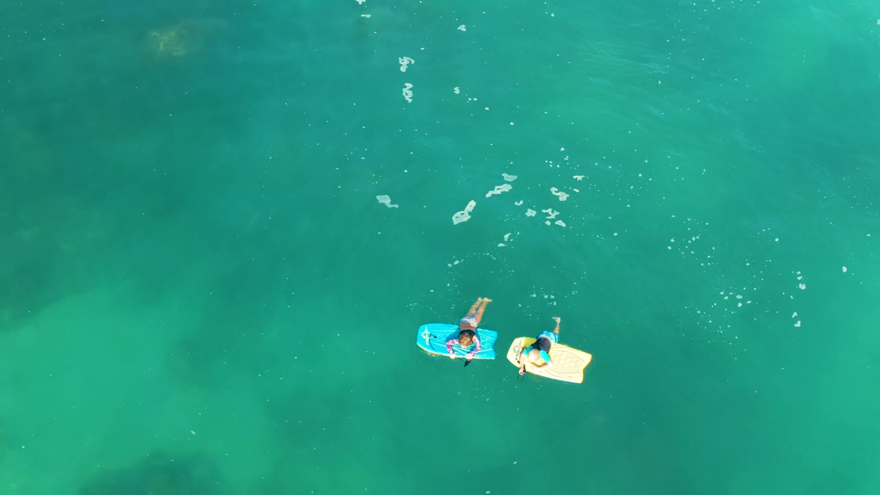 The two visible people drift slightly apart on their blue and beige boards in open turquoise water at Shipwrecks Los Cabos, with faint darker patches under the surface.