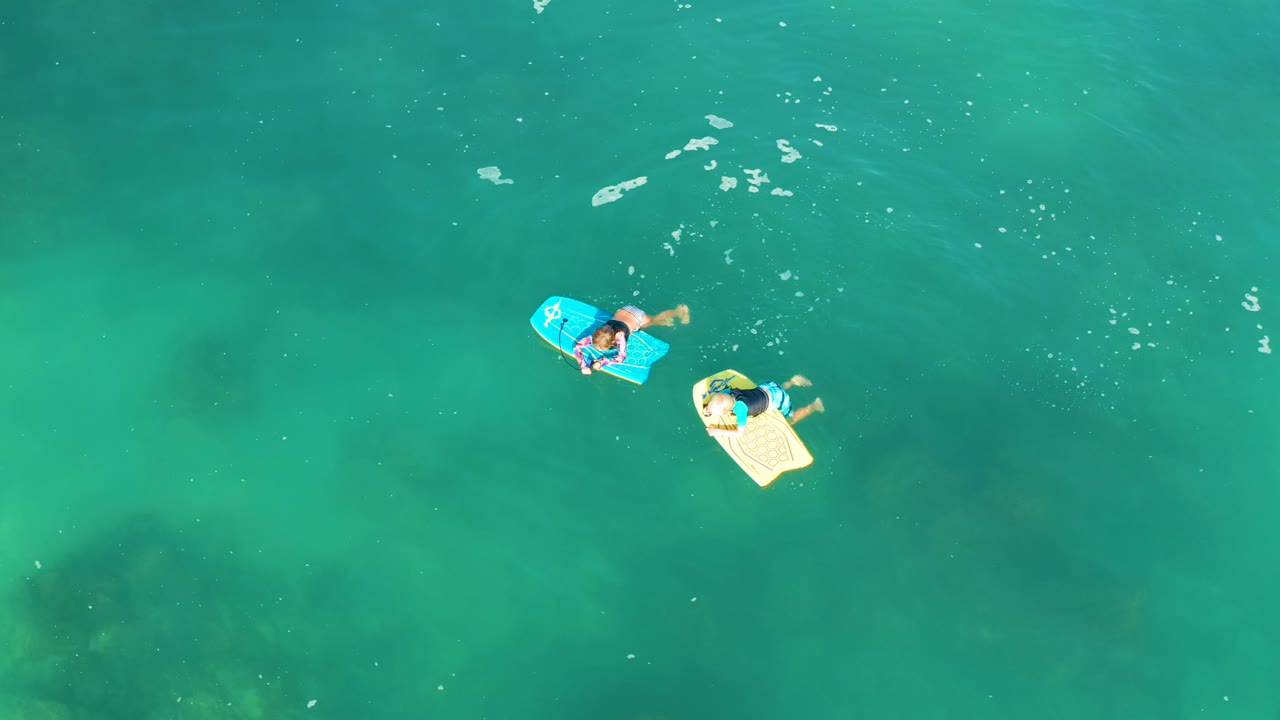 Overhead view at Shipwrecks Los Cabos showing two visible people lying on blue and pale yellow inflatable boards in clear turquoise water with small foam patches nearby.