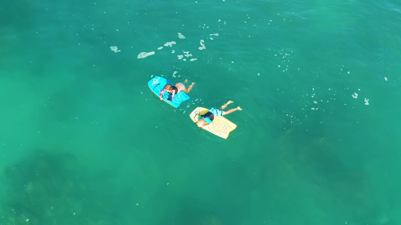 Overhead view at Shipwrecks Los Cabos of two people on blue and beige floating boards near the upper-left area of clear turquoise water.