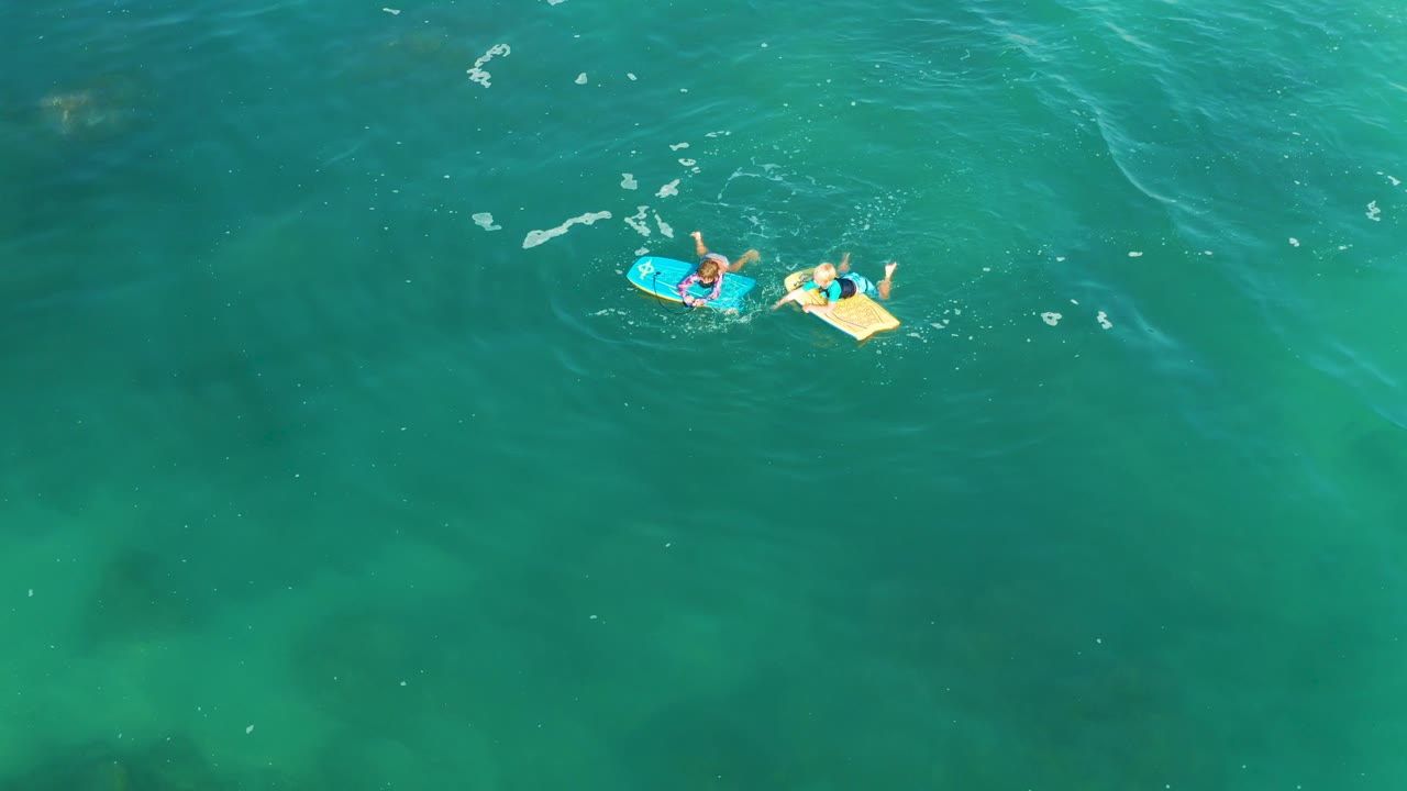 Overhead view at Shipwrecks Los Cabos of two visible people floating on blue and yellow bodyboards near the upper left of clear turquoise water with small foam streaks.