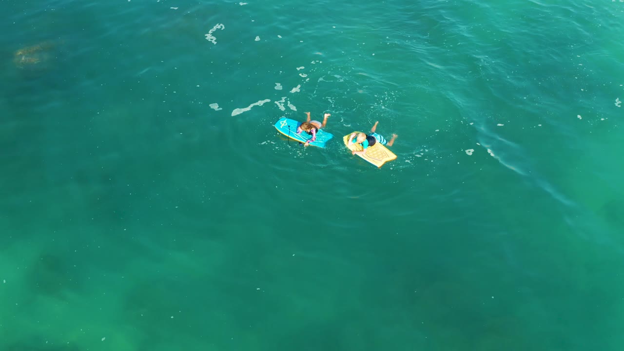 Aerial view over Shipwrecks Los Cabos water with two visible people on blue and yellow bodyboards near the top right, leaving most of the frame as open turquoise sea.