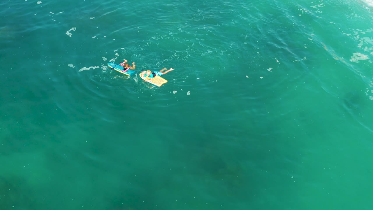 At Shipwrecks Los Cabos, two people float near the upper left of the frame on blue and yellow bodyboards in clear turquoise water with faint ripples.