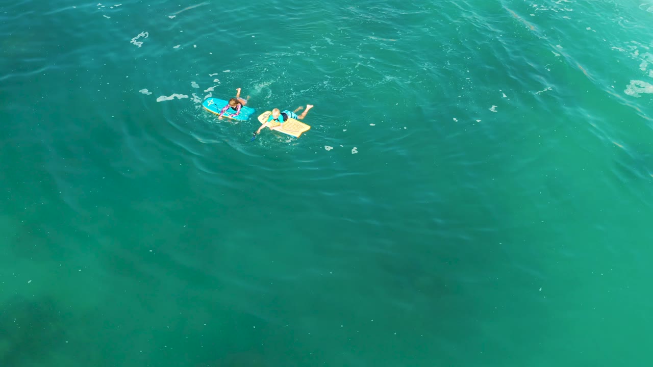 At Shipwrecks Los Cabos, the bodyboarders sit near the top center-right of the frame while circular ripples form around them in the turquoise sea.
