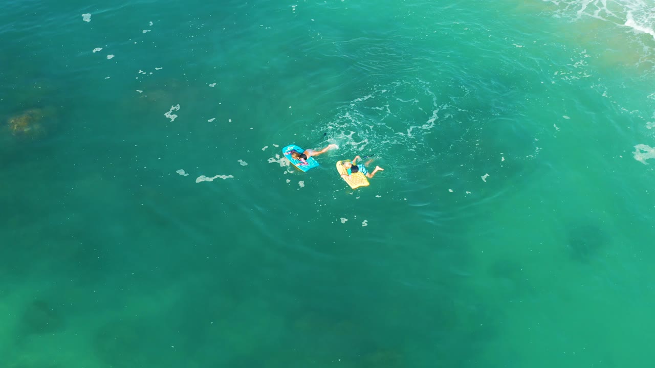 Overhead frame from Shipwrecks Los Cabos shows two people in clear turquoise water, one on a blue bodyboard and one on a light yellow float, with pale seabed patches below.