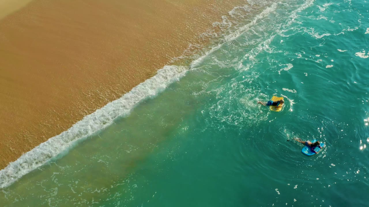 Aerial frame at Shipwrecks Los Cabos showing the yellow bodyboard closer to the upper right and the blue bodyboard lower right as whitewater thickens along the sand.