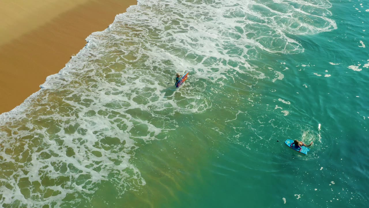 At Shipwrecks Los Cabos, the two distant bodyboarders hold separate positions as a band of white foam advances across the shallow water near the sand.