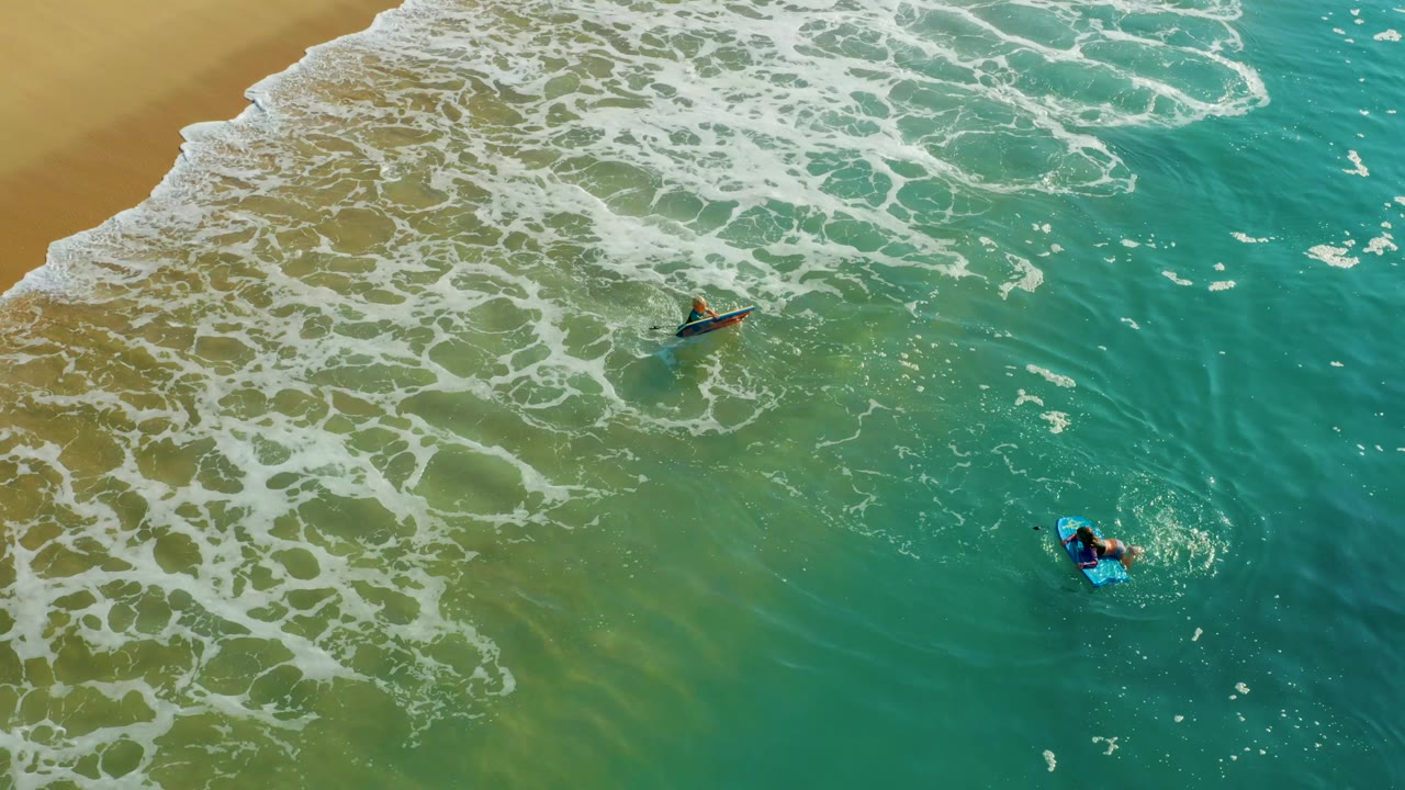At Shipwrecks Los Cabos, one bodyboarder is closer to the upper surf line while another stays lower in the turquoise water, with foam breaking across the sand side of the frame.