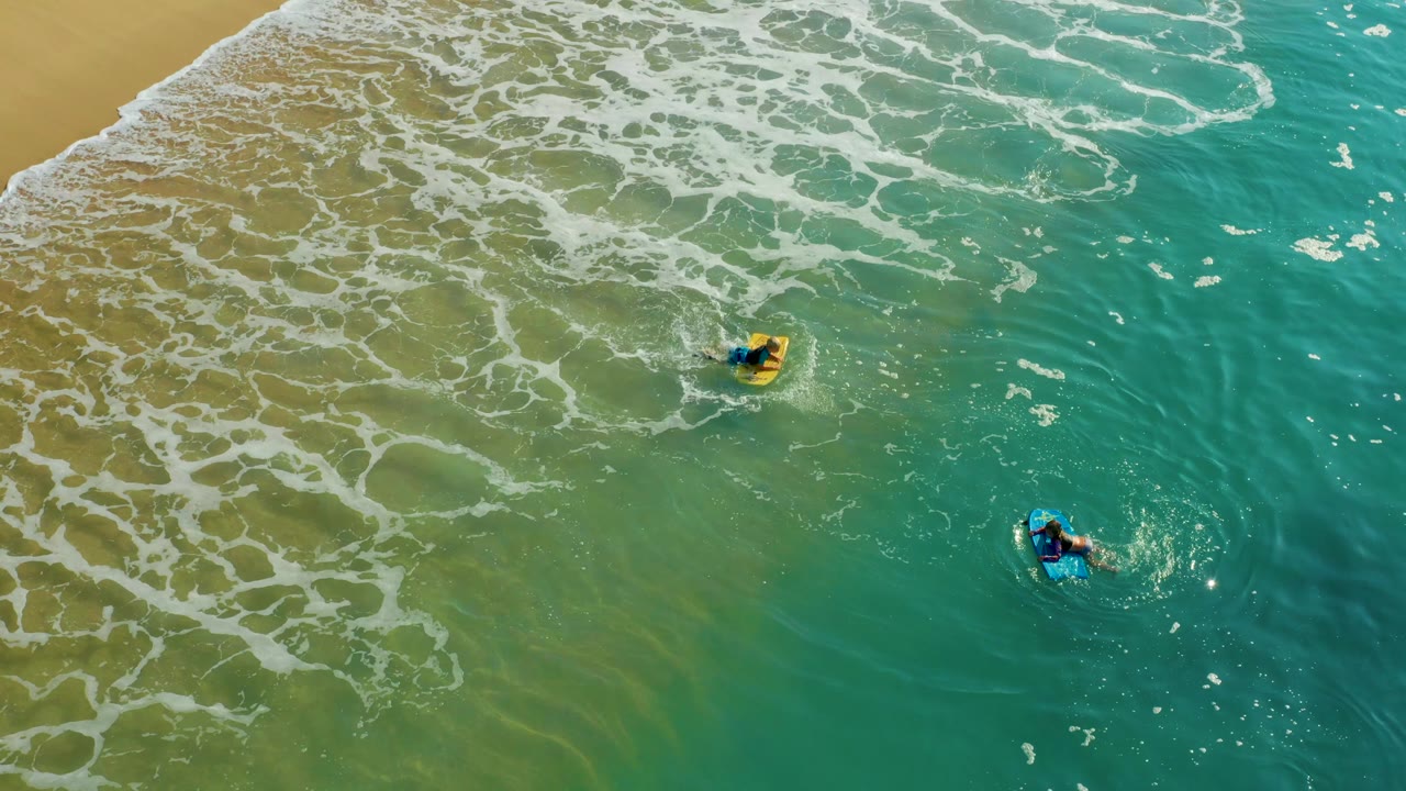 At Shipwrecks Los Cabos, a yellow-board bodyboarder moves near the center while a blue-board bodyboarder stays lower right, with foamy water spreading from the beach edge.