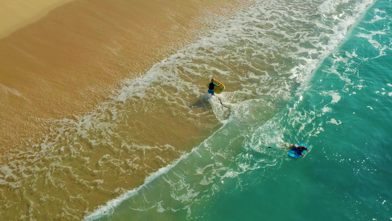 Overhead aerial frame at Shipwrecks Los Cabos shows two visible people with bodyboards separated by a diagonal line of white foam between wet sand and turquoise water.
