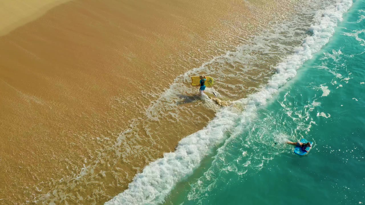 Aerial view at Shipwrecks Los Cabos shows one visible bodyboarder closer to the wet sand and another farther out in blue-green water as the whitewater thickens between them.