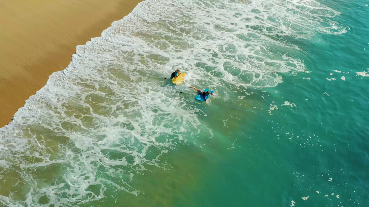 Overhead view at Shipwrecks Los Cabos showing two surfers on yellow and blue boards paddling below a foamy wave edge near tan sand.