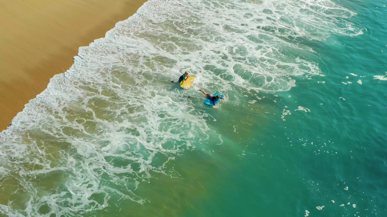 Aerial frame from Shipwrecks Los Cabos with the yellow-board surfer slightly above the blue-board surfer as foam spreads diagonally from the sandy beach.