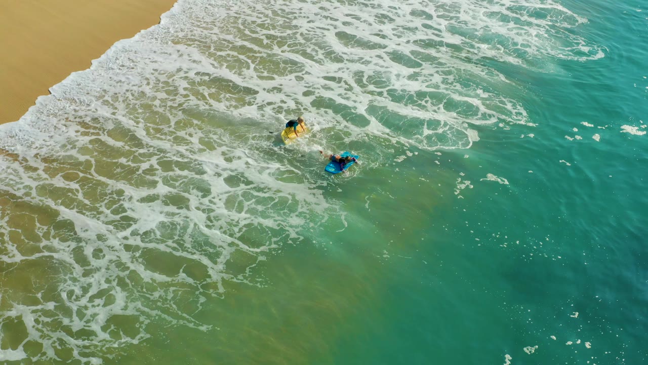 Overhead Shipwrecks Los Cabos view with two small surfers near the upper center, foam curling from the sand and clearer turquoise water filling the right side.