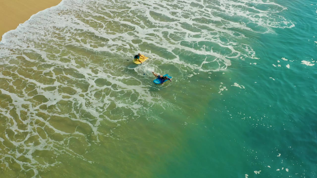 Two surfers paddling in green water beneath a broad sheet of foam.