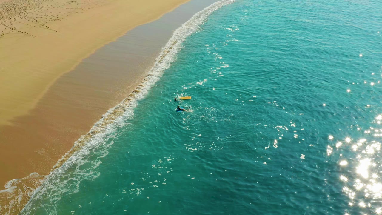 At Shipwrecks Los Cabos, two small bodyboarders paddle near a yellow board in clear turquoise water below a sandy beach with footprints and a thin white wave line.