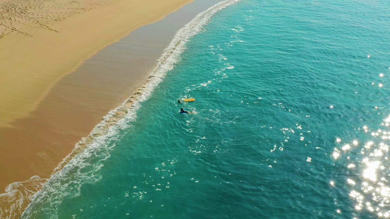 Yellow bodyboard and two paddlers near a diagonal shore break.