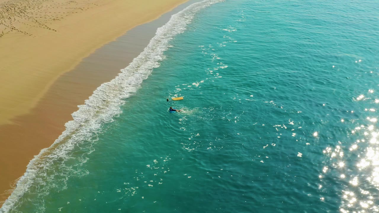 At Shipwrecks Los Cabos, two people remain offshore beside a yellow bodyboard while a wider white wave curls along the sandy beach above them.