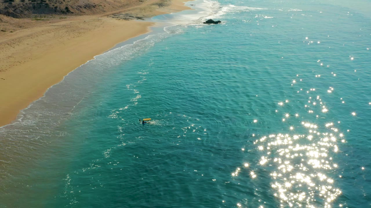 The surfer and yellow board remain offshore at Shipwrecks Los Cabos, with the foam line curving along the sandy beach and a dense cluster of water reflections on the right.