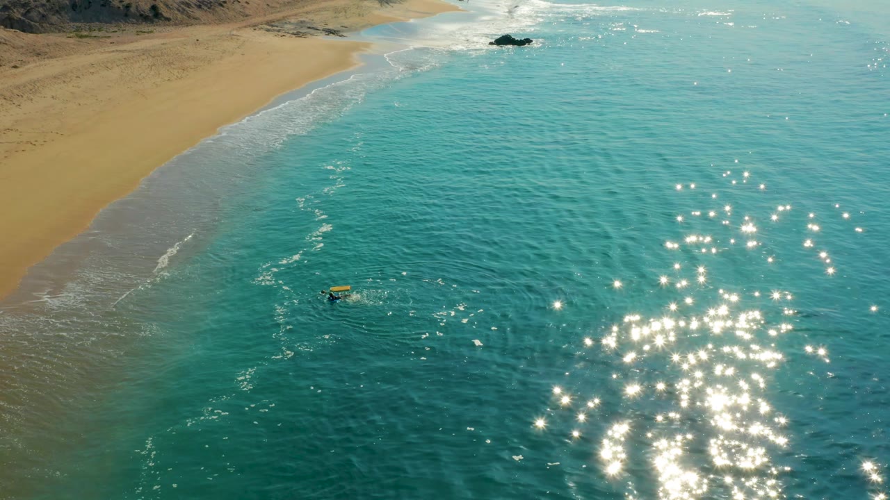 At the two-second mark, the yellow board sits slightly left of the main sparkle patch, with Shipwrecks Los Cabos sand and low surf visible along the upper-left shoreline.