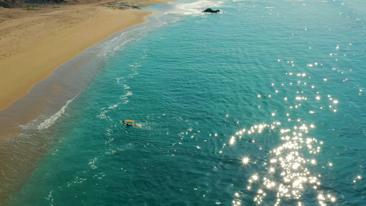 The surfer and yellow board appear closer to the lower-left water, with a broad sandy beach edge, a soft white break line, and bright reflections spread across the right side.