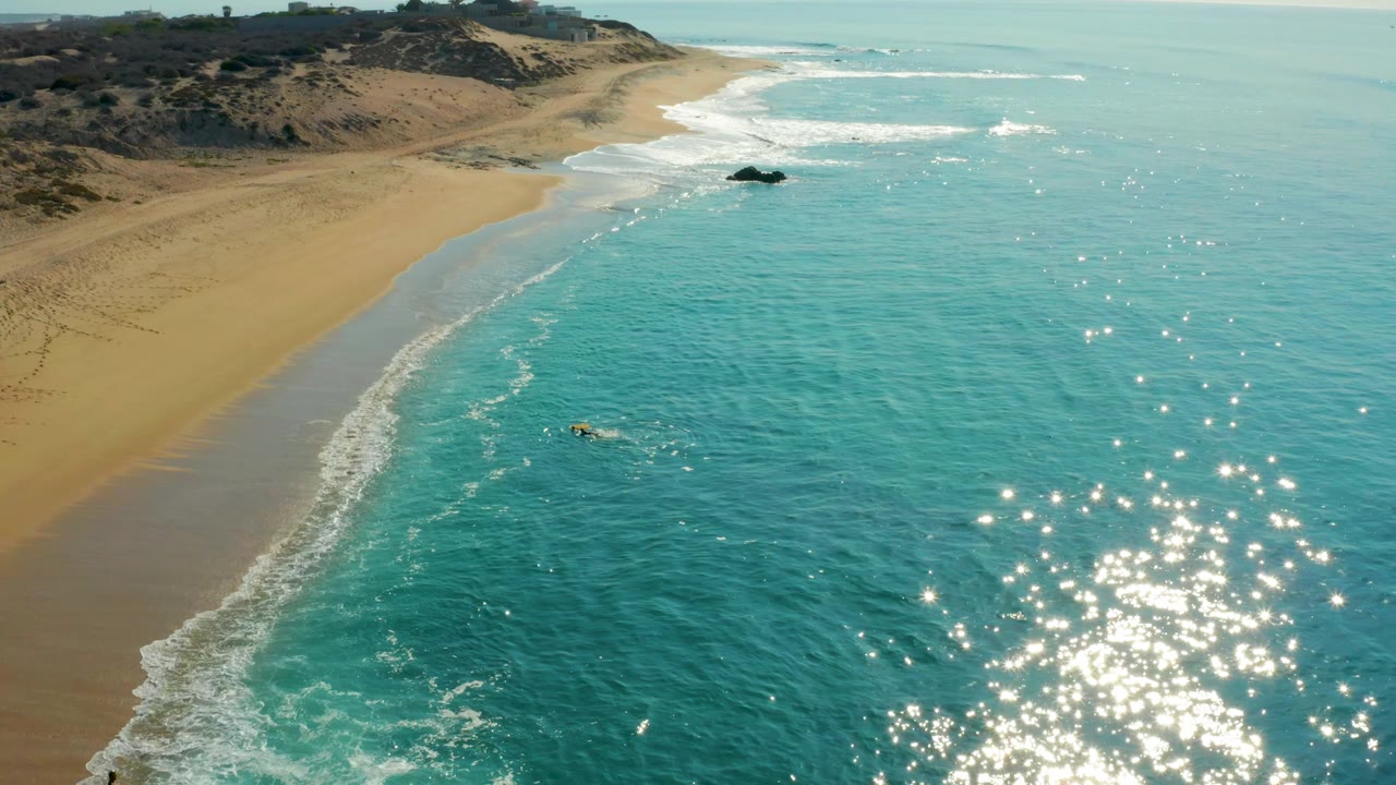 At 0:01, Shipwrecks Los Cabos appears from above with brighter sun glints on the right side of the ocean and the surfer still paddling below the diagonal surf line.