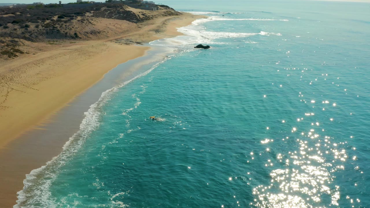 At 0:02, the aerial view shows the Shipwrecks Los Cabos beach curving into the distance, a dark rock near the upper surf, and the surfer positioned lower in the turquoise water.