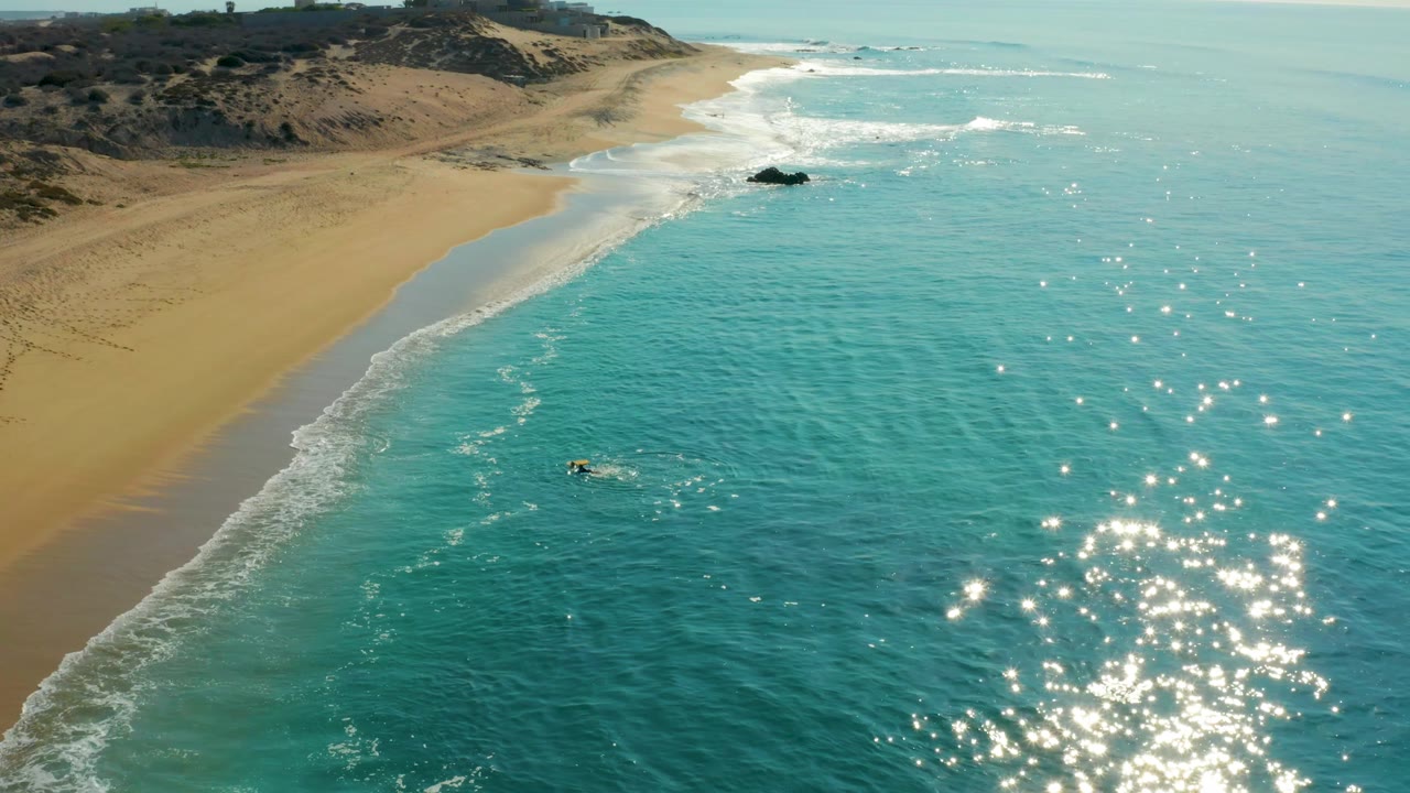 At 0:03, the Shipwrecks Los Cabos shoreline fills the left side, the turquoise ocean fills the right, and the surfer appears lower and closer to the sunlit water texture.