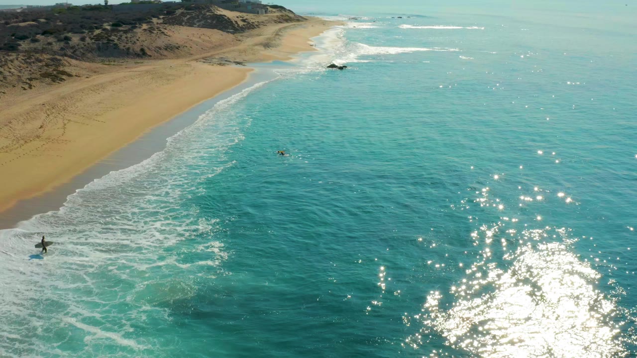 High coastal view with empty sand, blue-green water, surf foam, and surfers offshore.