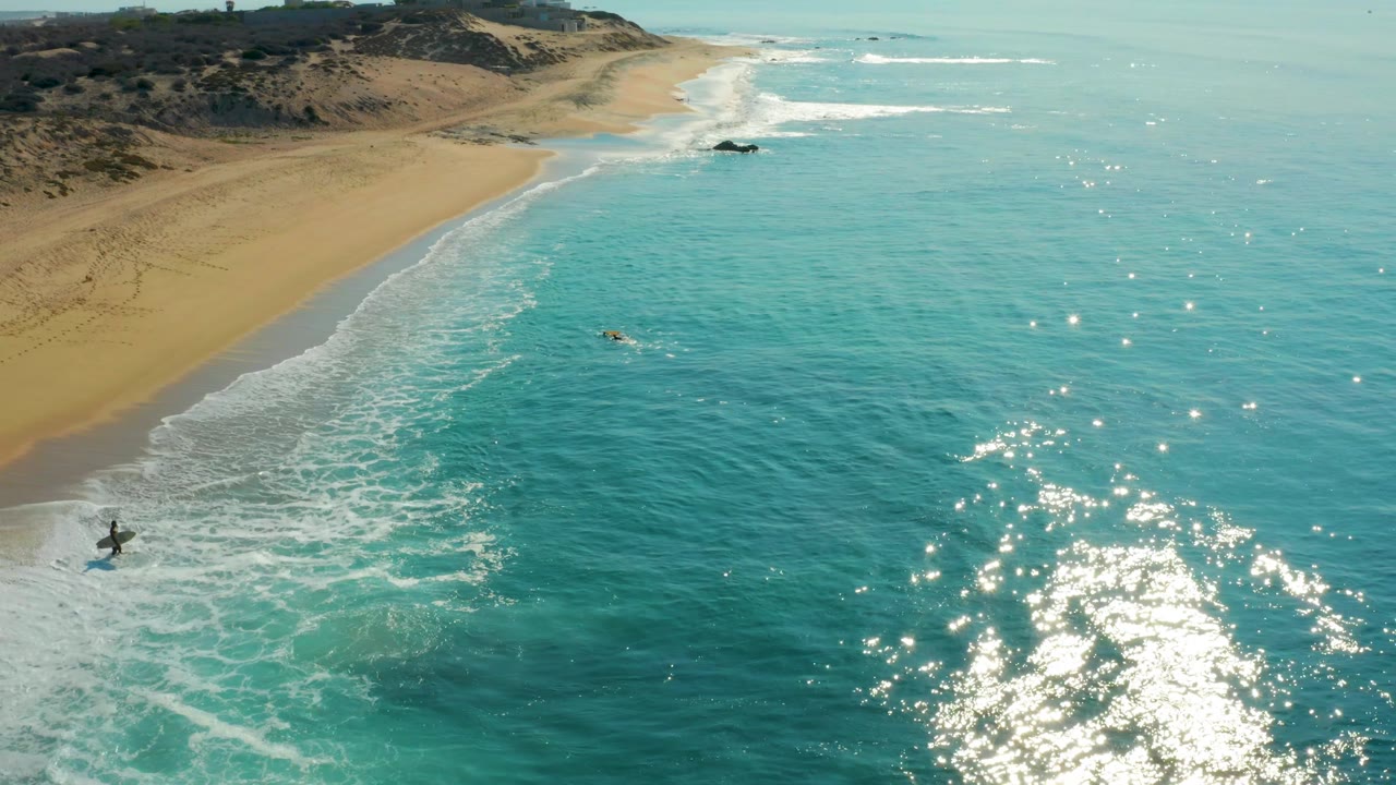 Aerial view at Shipwrecks Los Cabos with the shoreline curving through frame, sun glints clustered over the turquoise water, foamy surf reaching the sand, and small surfers visible offshore.