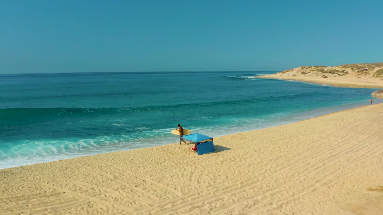 Person with surfboard near blue structure on open beach and ocean.