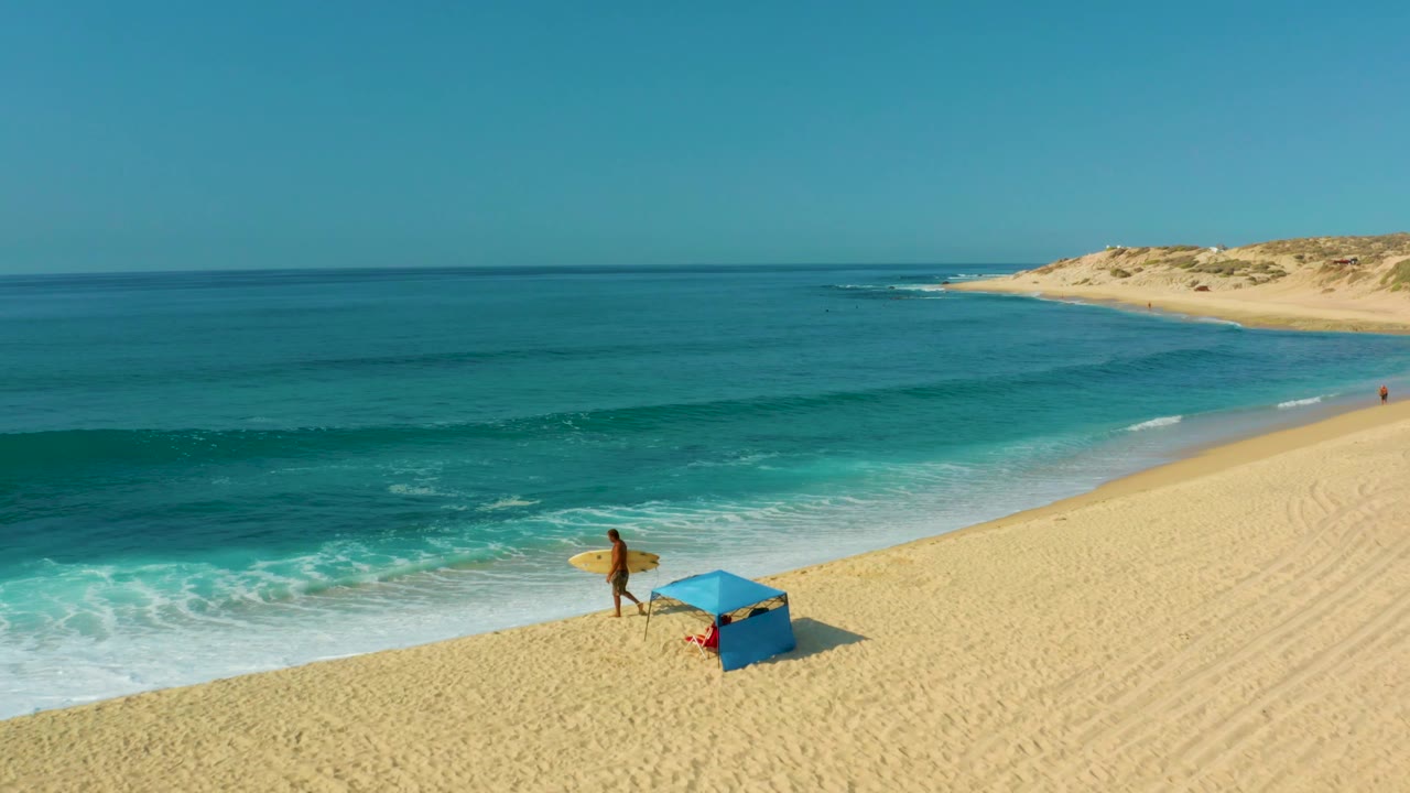 At Shipwrecks Los Cabos, the surfer has moved farther along the sand beside the blue umbrella, with white foam at the waterline and dunes in the background.