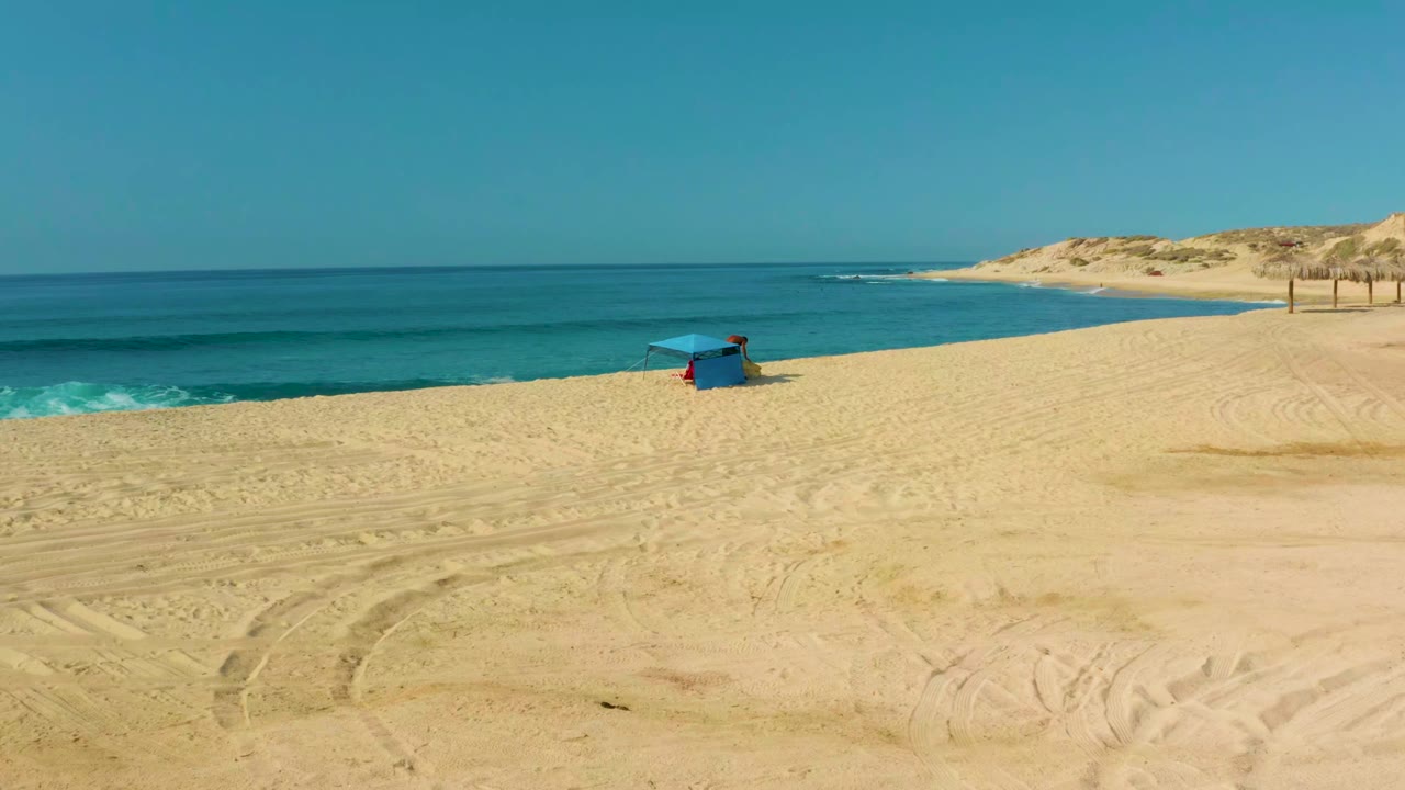 Wide view of Shipwrecks Los Cabos beach with pale sand tracks in the foreground, a blue hut near the waterline, turquoise ocean, and dunes in the distance.