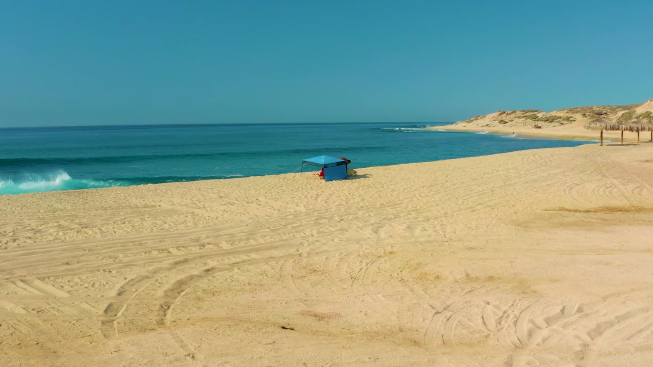 Bright sandy beach at Shipwrecks Los Cabos with a blue hut near the surf, rippled vehicle tracks across the foreground, and a low dune ridge beyond the shoreline.